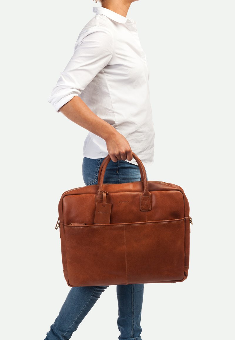 Borsa per laptop in pelle marrone con doppi manici, tasca frontale e etichetta per bagagli. Presenta una texture liscia e una forma strutturata.