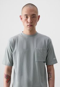 T-shirt gris à manches courtes en coton, avec une poche à l'avant, un col rond et des accents cousus. Texture lisse, coupe décontractée.