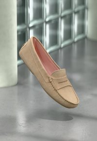Lys beige semskede loafer-sko med en glat overflade, syninger og en tekstureret sål, placeret i midten af luften mod en sløret baggrund.