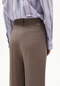 Braune maßgeschneiderte Hose mit glatter Textur, ausgestattet mit einer Gesäßtasche und Gürtelschlaufen. Kombiniert mit einem blau gestreiften Hemd.