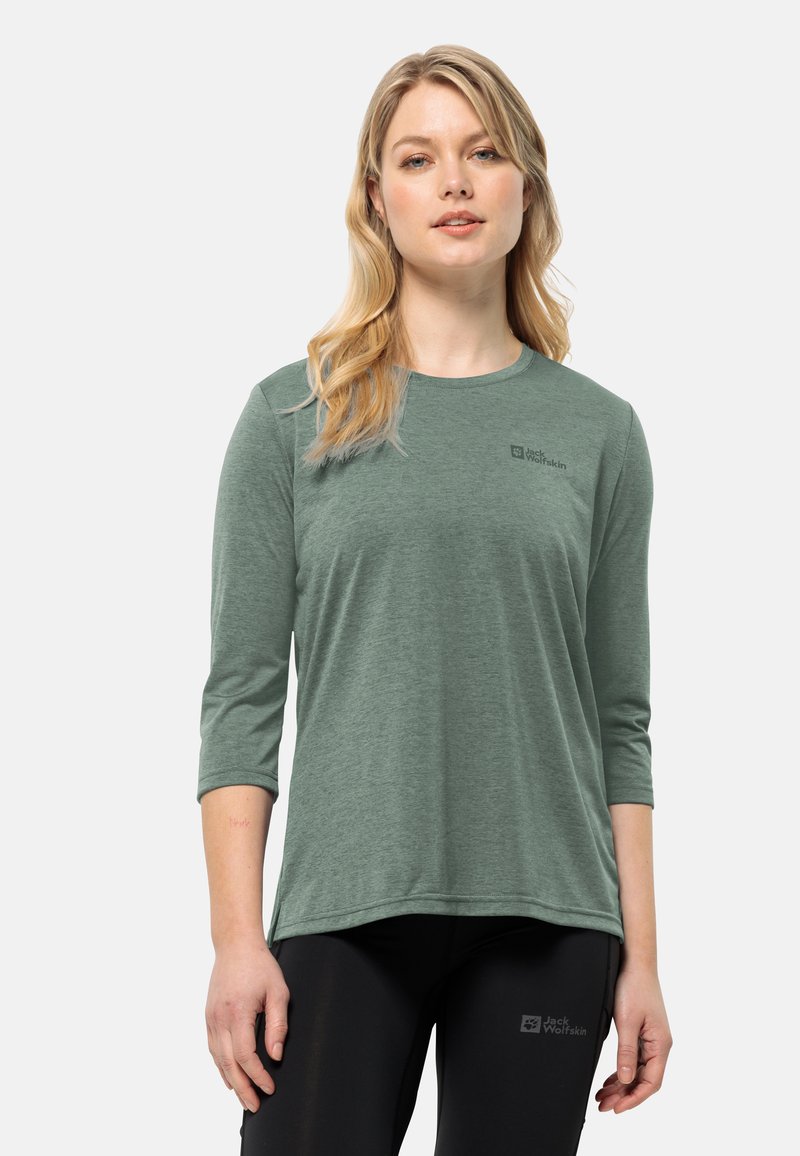 Donna bionda che indossa una maglia a maniche lunghe verde di Jack Wolfskin e pantaloni neri di Jack Wolfskin, in piedi con postura rilassata su uno sfondo bianco.