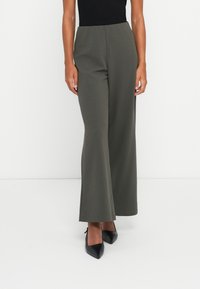 Frau in hoch taillierten, weiten olivgrünen Hosen, kombiniert mit schwarzen spitzen Absatzschuhen und einem schwarzen ärmellosen Top, stehend.