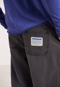 Čierne denimové nohavice majú zadné vrecko s modro-bielym štítkom, na ktorom je nápis "AMVDENIM". Textília má hladkú textúru a pevné prešívanie.