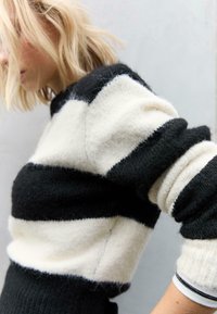 Pull rayé noir et blanc en matériau doux, avec un col rond et des manches retroussées. La texture semble duveteuse.