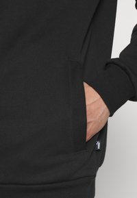 Black hoodie featuring a smooth fabric texture, side pockets, and ribbed cuffs. Small logo tag on pocket. Close-up view of hand in pocket.
