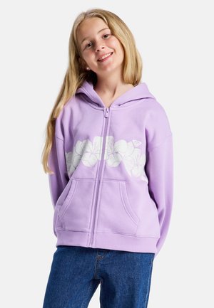 Lachend meisje met lang blond haar, gekleed in een lichtpaarse hoodie met ritssluiting en wit bloemmotief, en een blauwe spijkerbroek, tegen een effen achtergrond.