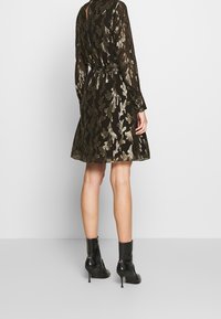 Robe noire avec un motif de feuilles dorées, manches longues, taille cintrée, jupe évasée et dos à découpe en forme de clé. Portée avec des bottines noires.
