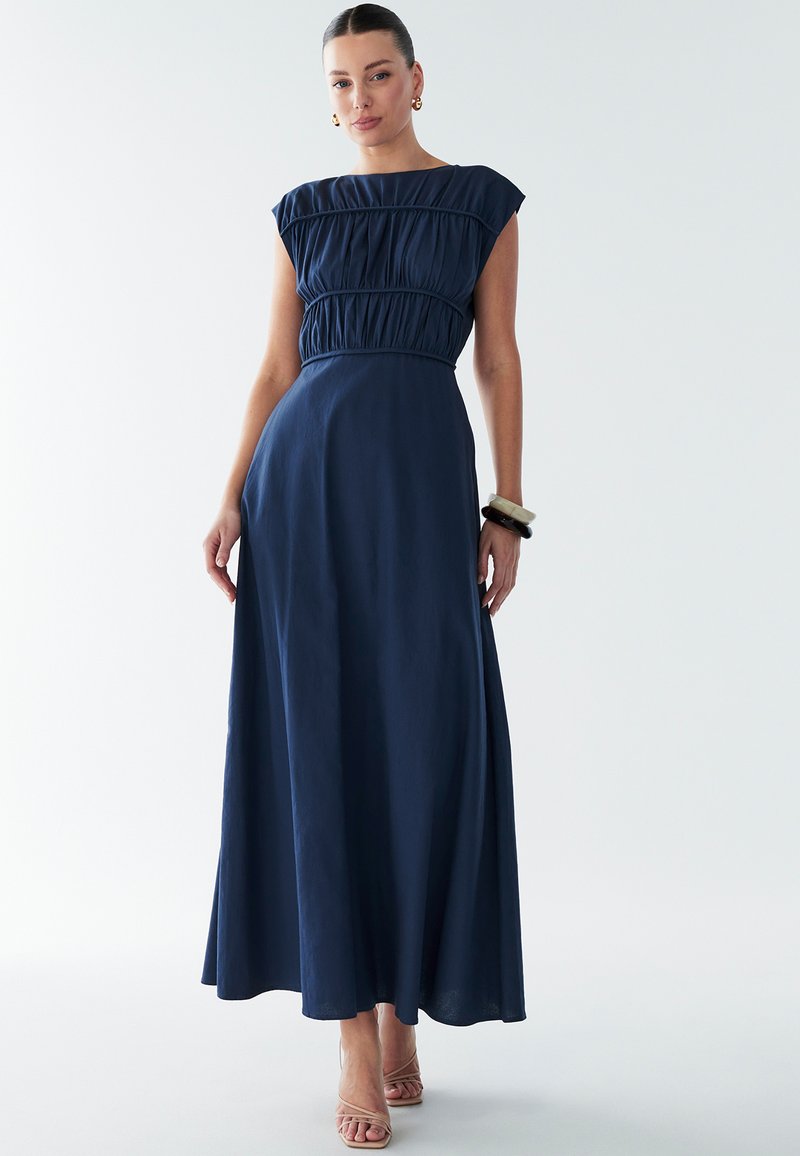 Tamsiai mėlyna maxi suknelė su surinktų raukšlių liemeniu ir trumpomis rankovėmis, lengvai krentanti medžiaga ir lygi tekstūra. Dėvima su skaidriomis aukštakulnėmis sandalais.
