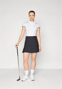 Άσπρο πόλο μπλουζάκι με κοντά μανίκια, μαύρο σορτς-φούστα και άσπρα αθλητικά παπούτσια. Η εμφάνιση είναι κατάλληλη για γκολφ και διαθέτει κομψό σχεδιασμό.