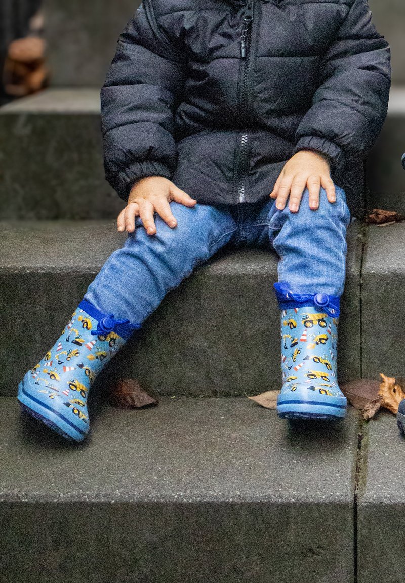 Kind zittend op betonnen trappen, gekleed in een blauwe spijkerbroek, een zwarte gewatteerde jas en blauwe regenlaarzen met patronen van bouwvoertuigen.