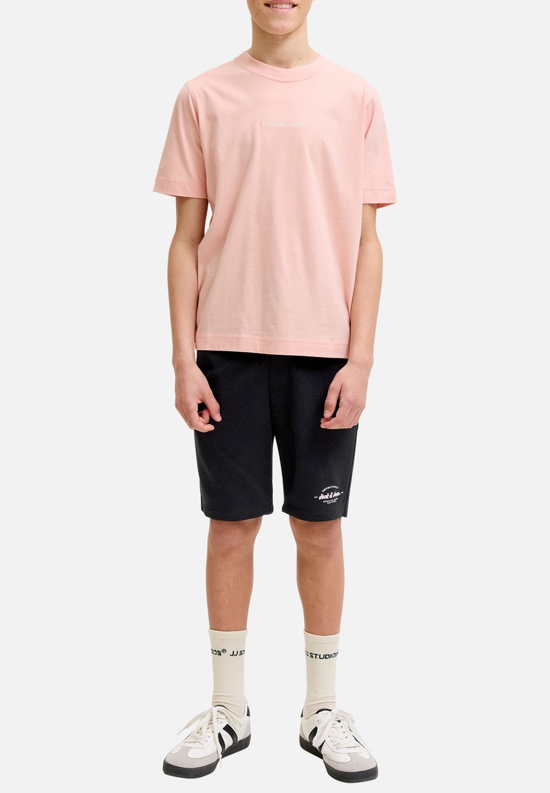 Adolescent portant un t-shirt rose clair à manches courtes, un short noir avec un texte blanc, des chaussettes crème et des baskets blanches, debout sur un fond blanc.