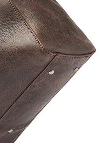 Esquina de bolsa de cuero marrón con superficie texturizada, costuras visibles y tachuelas metálicas. La forma es angular, mostrando un material duradero.