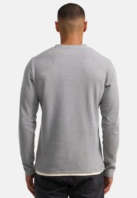 Camiseta gris de manga larga con un patrón texturizado de tejido tipo gofre. Presenta un cuello redondo y puños acanalados, llevada sobre una camisa blanca en el dobladillo.