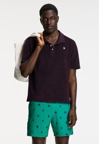 Short-sleeve, dark purple terry polo with three buttons, worn with green swim trunks featuring navy bird silhouettes. Cotton tote over shoulder.