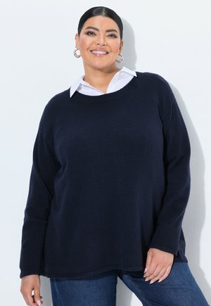 Lachende vrouw met donker haar in een marineblauwe trui over een wit overhemd met kraag, met hoepeloorbellen, poserend tegen een lichte achtergrond.