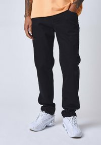 Pantalones de mezclilla negros con corte recto, que cuentan con presillas para cinturón y bolsillos laterales, combinados con zapatillas deportivas blancas y una camiseta naranja.