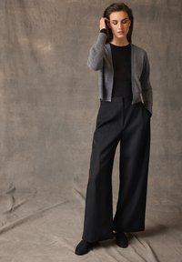 Un cardigan en tricot gris sur un haut noir associé à un pantalon noir taille haute. Le tissu semble doux et le pantalon a une coupe ample et ajustée.