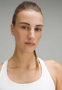 Close-up of a person with medium skin tone, wearing a white tank top. Hair is slicked back with a small braid, neutral background.