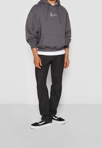 Sudadera con capucha gris, con bolsillo frontal y logo blanco, sobre una camiseta blanca, combinada con jeans oscuros y zapatillas negras.