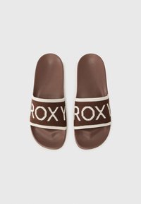 Smeđe sandale na petu s bijelim pletenim trakama koje sadrže tekst "ROXY", ravni potplat i glatku završnicu.