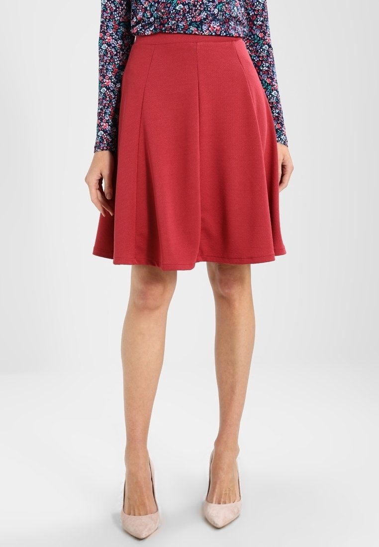 Roter, ausgestellter Rock aus glattem Stoff, mit Falten an der Taille und knielanger Schnittführung. Kombiniert mit hellen, spitzen Schuhen.