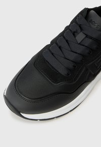 Scarpa sportiva nera con tomaia in tessuto e materiale sintetico, dotata di lacci neri piatti, colletto imbottito e suola in gomma bianca.