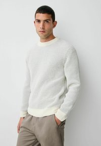 Pulover alb din tricot cu un model texturat, guler rotund cu nervuri și manșete. Purtați cu pantaloni de culoare deschisă.