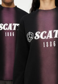 Δύο άτομα φορούν μαύρα και μοβ φούτερ με διαβάθμιση χρώματος, με λευκό κείμενο "SCAT" και "1886" και ένα γεωμετρικό λογότυπο στο στήθος.