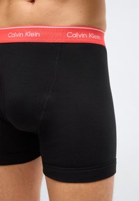 Κοντινό πλάνο ενός ατόμου που φοράει μαύρα άνδρικα εσώρουχα boxer με κόκκινη μέση που φέρει το λογότυπο Calvin Klein σε απλό φόντο.