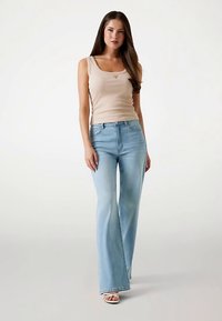 Beiges Tanktop und hellblaue ausgestellte Jeans. Das Tanktop hat einen rund ausgeschnittenen Ausschnitt, und die Jeans verfügen über eine hohe Taille und gerade Beinform.