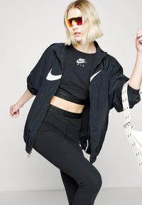 Črna vetrovka z belim Nike logotipom, črn kratki zgornji del z logotipom, črne legice, rdeče in rumene zrcalne sončne očala, bela torbica s študnimi detajli.