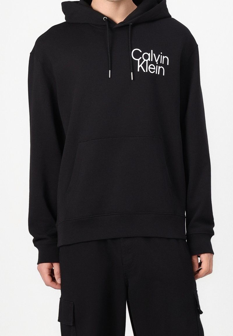 Personne portant un sweat à capuche Calvin Klein noir avec poche avant et logo blanc sur le haut gauche de la poitrine, assorti à un pantalon noir avec poches latérales.