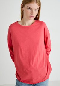 Jonge vrouw met lichte huid, steil rood haar, gekleed in grote oorringen, een rode langwerpige blouse en lichtblauwe spijkerbroek, tegen een effen achtergrond.