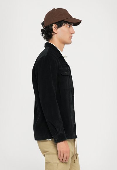 Black corduroy shirt with front pockets and button closure, paired with a brown cap. Model wears beige cargo pants. Side profile view.