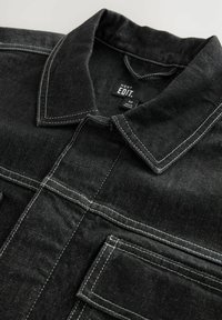 Giacca di jeans nera con cuciture bianche a contrasto, colletto e tasche sul petto, con un aspetto di tessuto testurizzato.