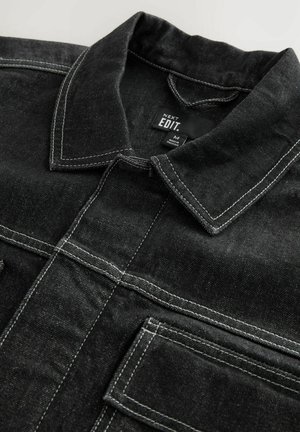 Chaqueta de mezclilla negra con costuras blancas en contraste, cuello tipo solapa y bolsillos en el pecho, con un acabado de tela texturizada.