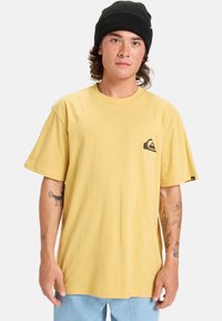 Žluté bavlněné tričko volného střihu s malým černým logem na hrudi. Nosit s světle modrými džínsovými šortkami a černou čepicí.