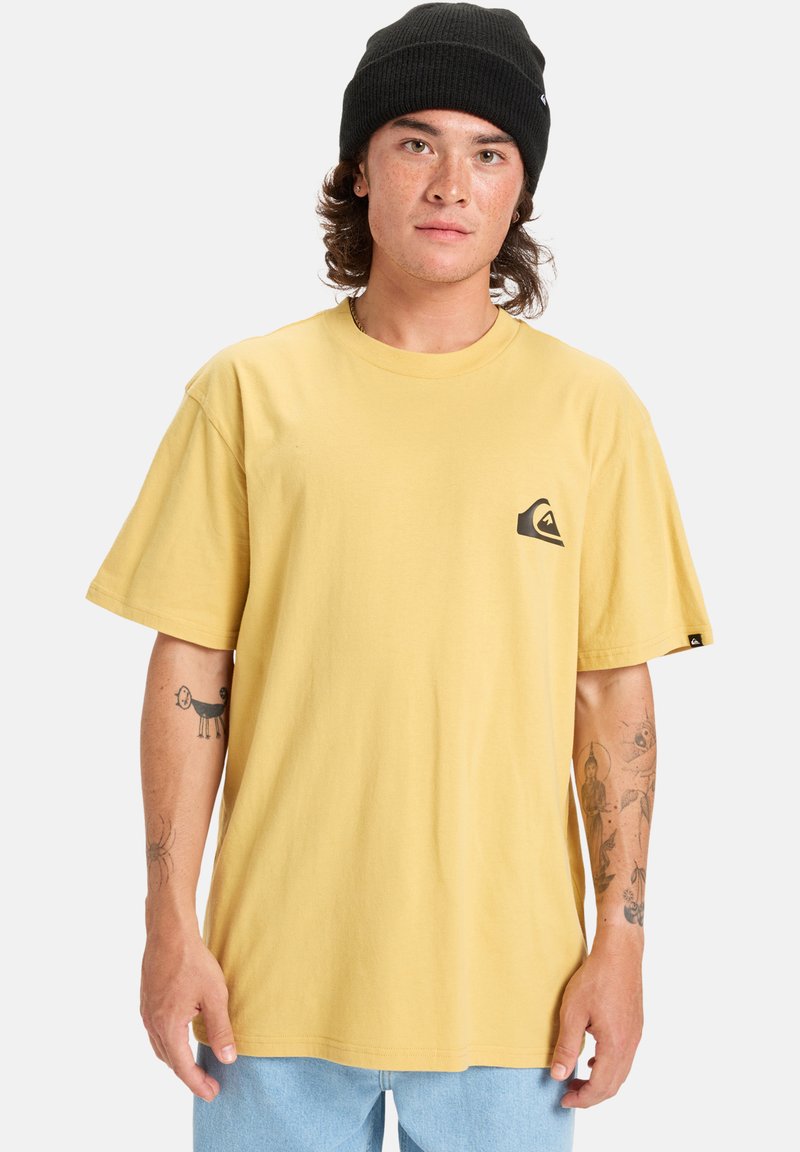 Žluté bavlněné tričko volného střihu s malým černým logem na hrudi. Nosit s světle modrými džínsovými šortkami a černou čepicí.