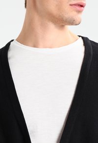 Närbild av en mans nacke och nederdel av ansiktet, iklädd en vit t-shirt med rund hals, lager på lager med en svart öppen cardigan.