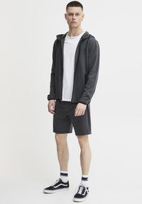 Grijze rits hoodie met zwarte accenten, gecombineerd met bijpassende grijze shorts. Draagt een wit T-shirt, zwarte sneakers en gestreepte sokken.