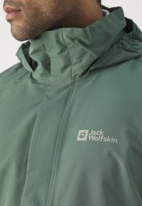 Veste imperméable verte avec capuche, au toucher lisse et avec un logo sur la poitrine. Fermeture éclair et col réglable.