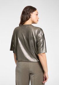 Metaliczna, krótka bluzka z krótkim rękawem o luźnym kroju, z błyszczącą, brązową powierzchnią i prostym dołem, zestawiona z miękkimi spodniami w neutralnych odcieniach.