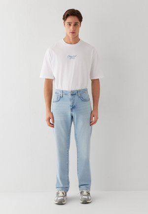 Junger Mann steht, trägt ein weißes T-Shirt mit dem Schriftzug „Coastal zone“, hellblaue Jeans und graue Sneaker vor weißem Hintergrund.