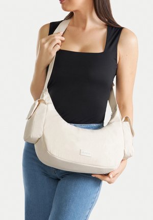 Frau trägt schwarzes ärmelloses Oberteil und blaue Jeans und hält große beige Umhängetasche mit vorderen Taschen und breitem Träger.