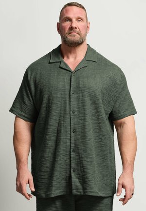 Szeroki mężczyzna z brodą stoi zwrócony przodem, ubrany w teksturowaną ciemnozieloną koszulę z krótkim rękawem na guziki oraz pasujące spodnie.