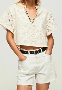 Top court de couleur crème avec des motifs en œillets et un col ondulé, associé à un short blanc taille haute et une ceinture noire.