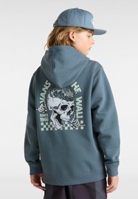 Sudadera gris con un gráfico de calavera y un patrón de tablero de ajedrez en la parte posterior; cuenta con un bolsillo frontal y capucha ajustable. La gorra es de un azul a juego.