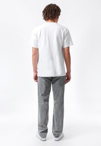 Mann mit lockigem Haar, der mit dem Rücken zur Kamera steht, trägt ein schlichtes weißes T-Shirt, graue Jeans und weiße Turnschuhe vor weißem Hintergrund.