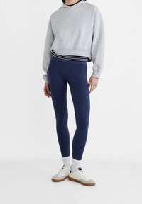 Sudadera recortada gris claro con capucha, leggings acanalados azul marino y zapatillas blancas con suelas marrones. Capa con una camiseta de rayas debajo.