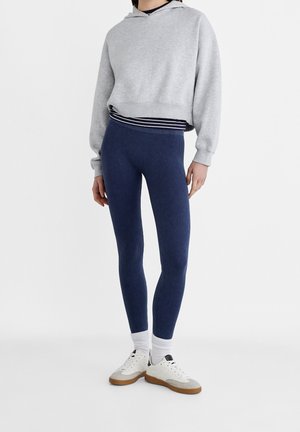 Vaaleanharmaa lyhyt huppari hupulla, tummansiniset ribbileggingsit ja valkoiset lenkkarit ruskeilla pohjilla. Kerroksittain raidallinen paita alla.